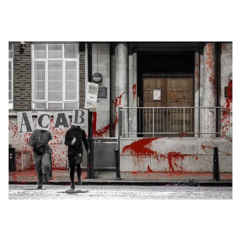 Two people stand in front of a paint splattered cop shop their faces obscured Text reads: ACAB