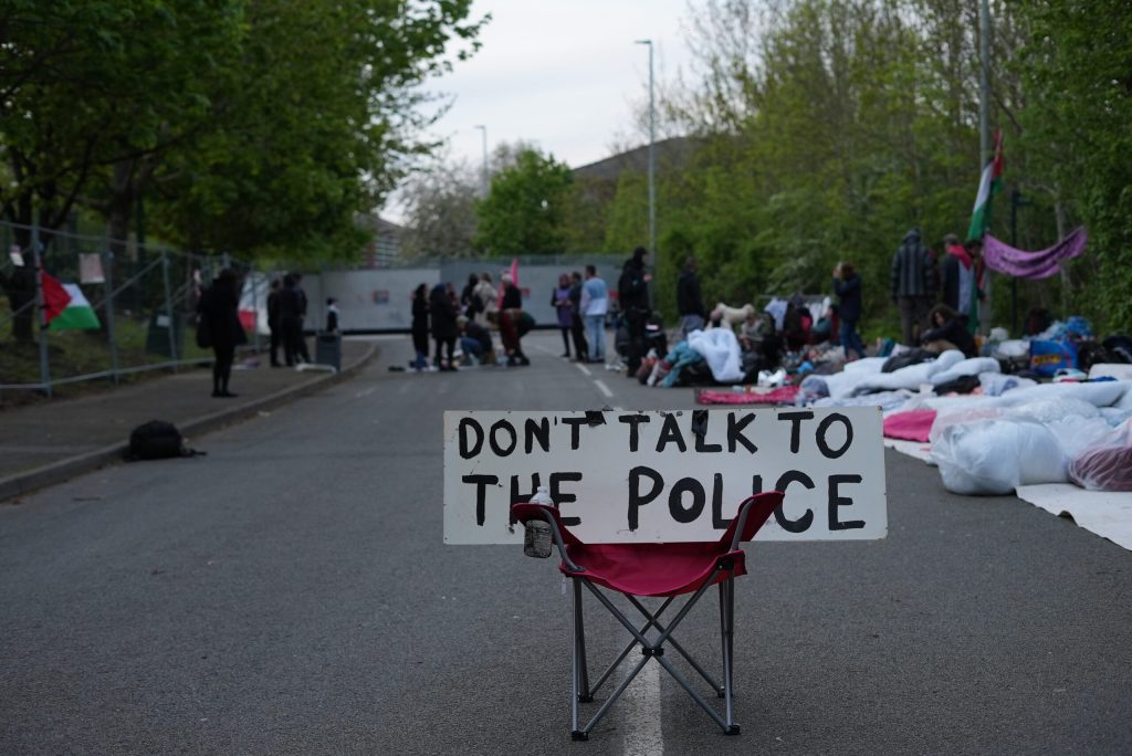 A chair with a sign reading DON'T TALK TO THE POLICE is the the foreground with people in the background