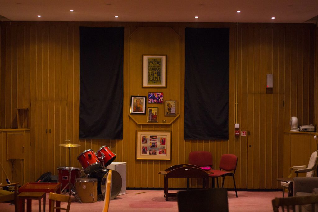 [ID: interior of a large warmly lit room with chairs and a drum kit]