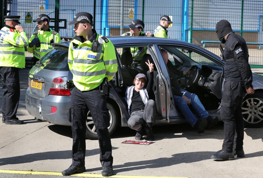 A person is locked on in a car and gives the peace symbol while surrounded by cops outside the gates of Elbit's weapons factory
