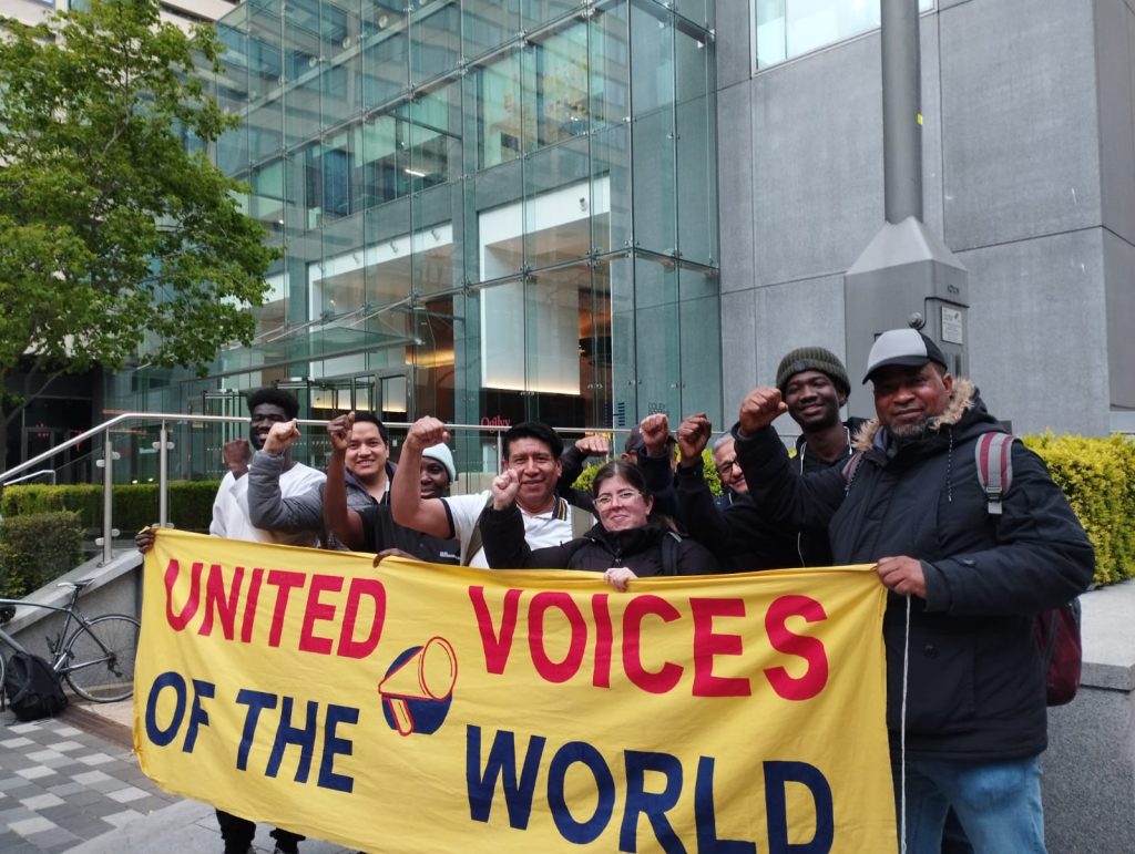People stand with with fists in the air behind a banner that reads UNITED VOICES OF THE WORLD