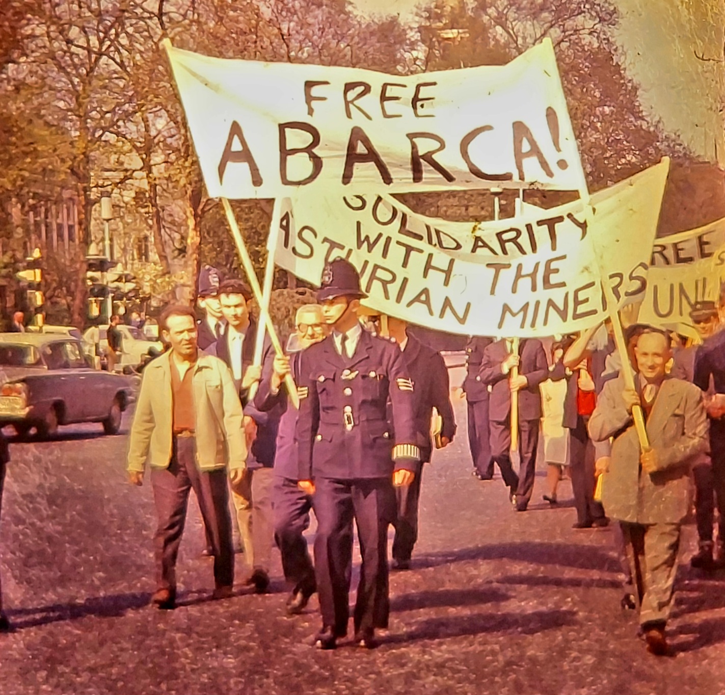 Monochrome photo of demonstration on a British street showing men holding banners reading Free Abarca! and Solidarity with the Asturian miners. A british police officer is walking at front with one of the demonstrators looking ascanse at him