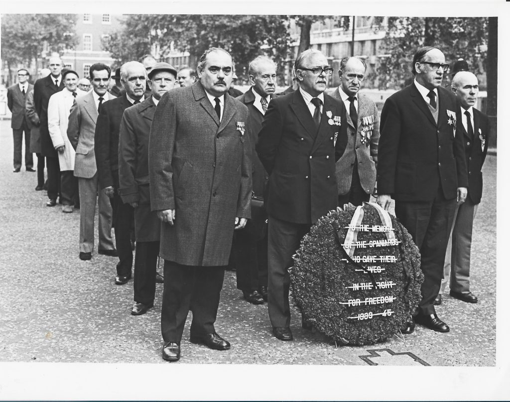 Black and white photo of column of older men wearing civilian clothes and medals with a large wreath at the front of the column, reading to the memory of the spaniards who gave their lives in the fight for freedom 1939-45