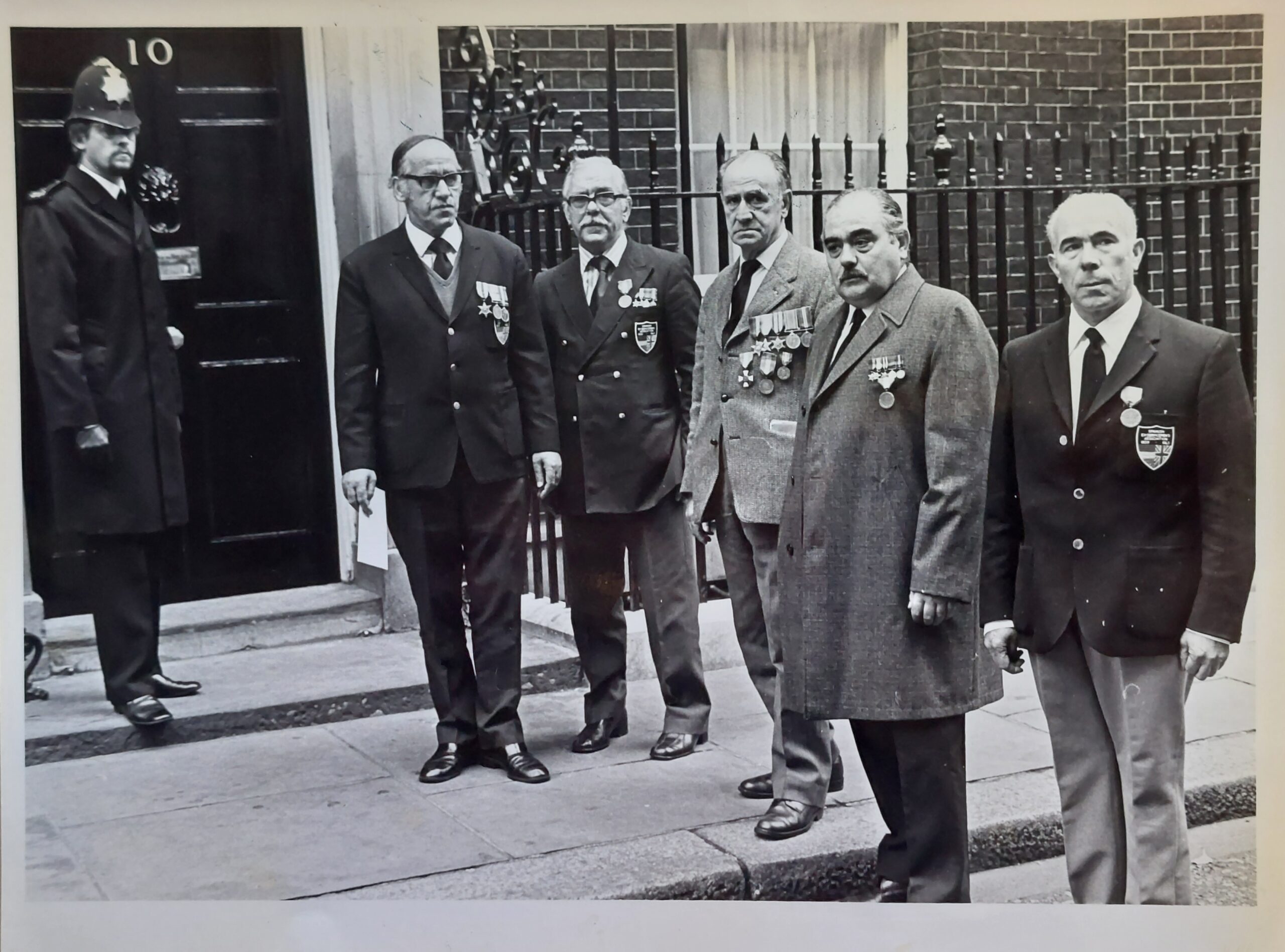 Black and white photo of 10 downing street showing five older men in civilian clothes and medals plus a police officer